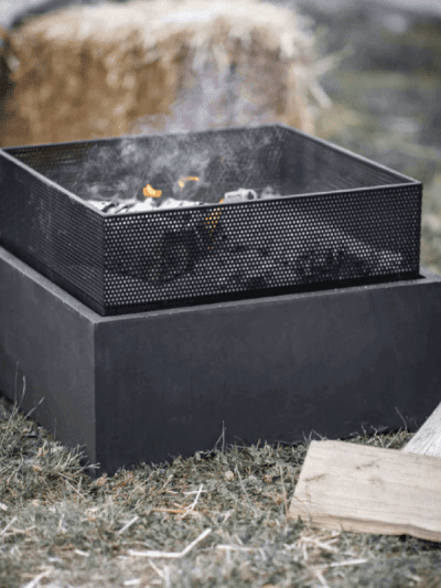 Square steel garden fire pit alight, next to pile of logs in a garden