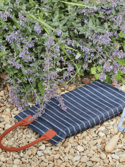 Sophie Conran Garden Kneeler, navy and white striped with a tan leather handle on beige gravel next to plants