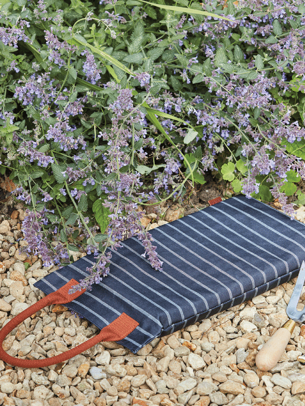Sophie Conran Garden Kneeler, navy and white striped with a tan leather handle on beige gravel next to plants