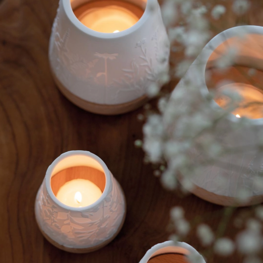 White unglazed porcelain tealight holder with embossed flowers on an oiled wooden base by Räder, casting a warm glow when lit.