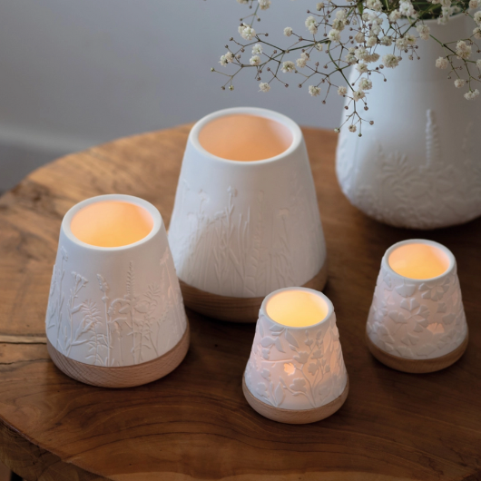 White unglazed porcelain tealight holder with embossed flowers on an oiled wooden base by Räder, casting a warm glow when lit.