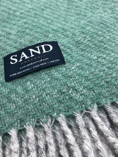 Sand Cornwall sea-green pure new wool blanket with cream tassels and soft textured weave, neatly folded on a sofa.