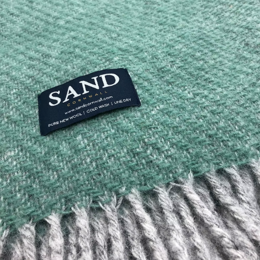 Sand Cornwall sea-green pure new wool blanket with cream tassels and soft textured weave, neatly folded on a sofa.