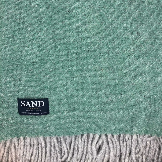 Sand Cornwall sea-green pure new wool blanket with cream tassels and soft textured weave, neatly folded on a sofa.