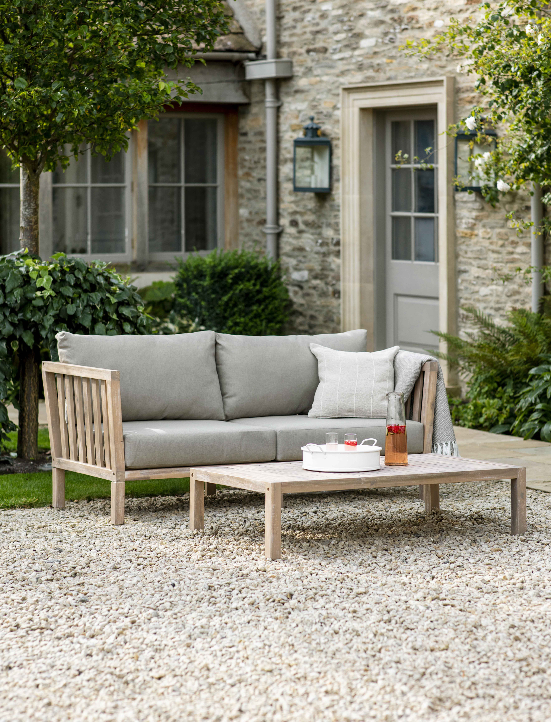 Garden Trading Porthallow 2 seater sofa in natural acacia wood with grey cushions styled on an outdoor patio.